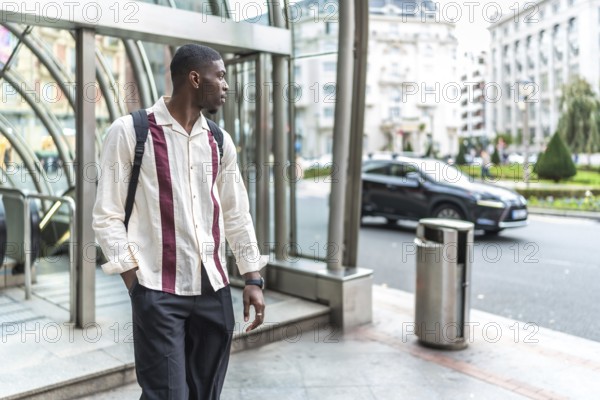 Young black man carrying a backpack and wearing a stylish shirt, standing confidently on a city street, looking away while waiting for public transportation from an urban subway station