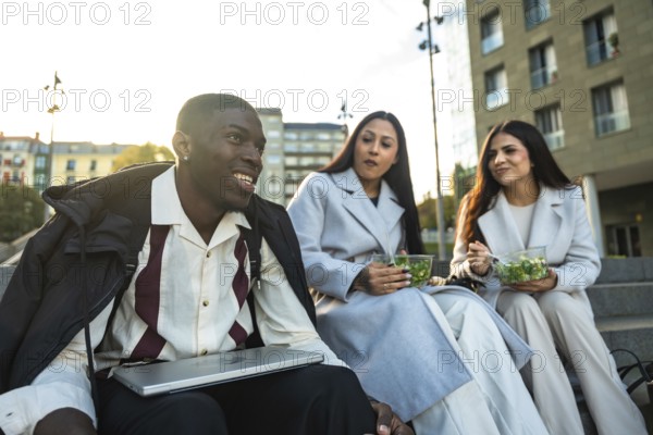 Diverse group of young adult colleagues or friends smiling and having a relaxed conversation while eating healthy salads on stairs during their outdoor lunch break in a city setting