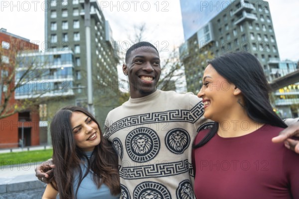 Diverse group of three young friends walking together on a city street, one man embracing two women, all happy and smiling while enjoying friendship and urban life