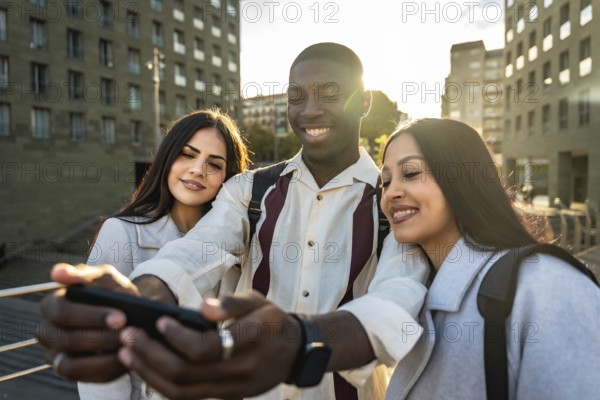 Diverse group of young friends is smiling and laughing together while taking a selfie with a smartphone outdoors in a city, sharing a happy moment of friendship and connection