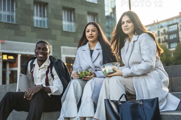 Diverse colleagues sitting on steps and smiling while enjoying healthy salads during a casual urban lunch break, representing modern work life balance and friendship