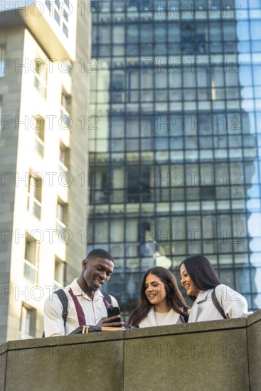 Three diverse students, a man and two women, smiling and looking at a smartphone, sharing content and interacting socially outdoors in a modern city environment