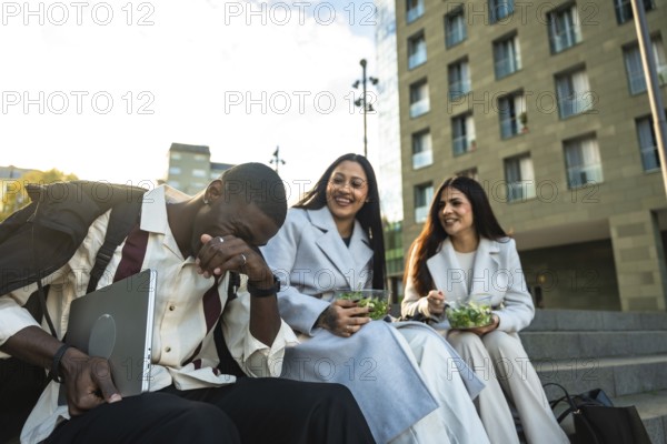 Diverse colleagues enjoying their lunch break outdoors, sitting on stairs in the city, laughing and eating salads while sharing a happy moment together