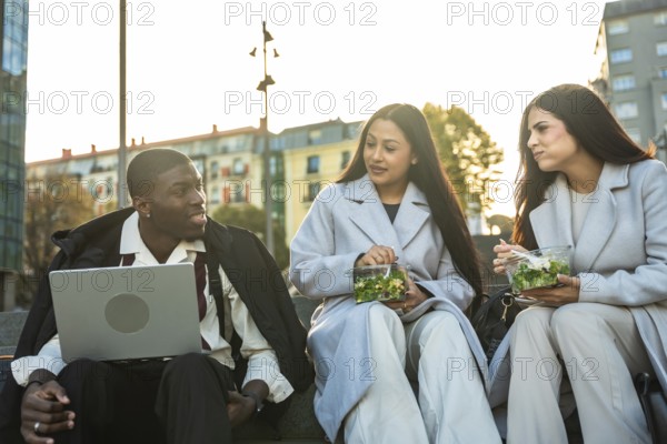 Diverse business colleagues enjoying a relaxed lunch break outdoors in the city, sitting on steps, eating salads, and engaging in conversation with a laptop nearby