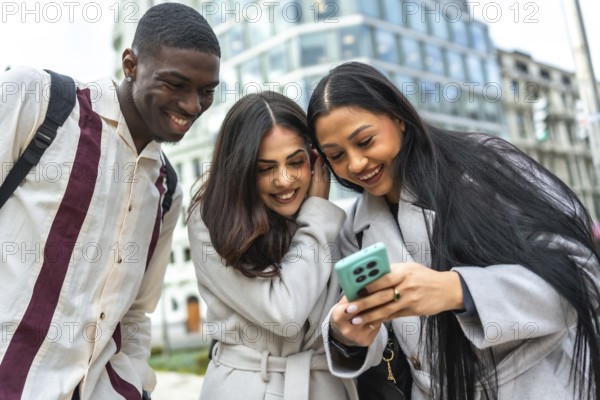Diverse group of young adult friends smiling and laughing while watching something on a smartphone together, sharing a happy and connected moment in a modern city environment