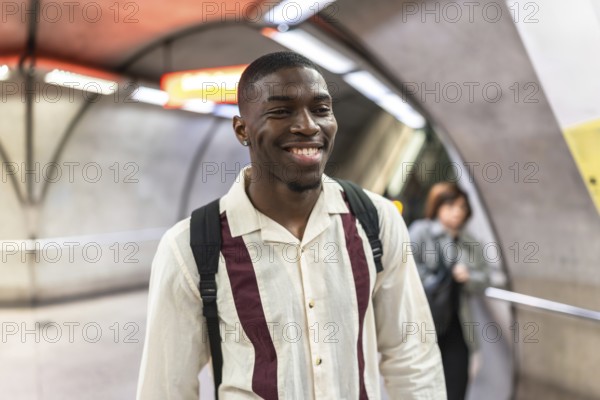 Smiling young african american man with a backpack walking through a modern underground subway station, enjoying his daily commute with a happy and positive expression