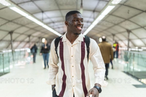 Young black man with backpack walking through bright modern airport walkway, smiling confidently as he commutes or travels, embodying freedom, progress and a positive urban journey