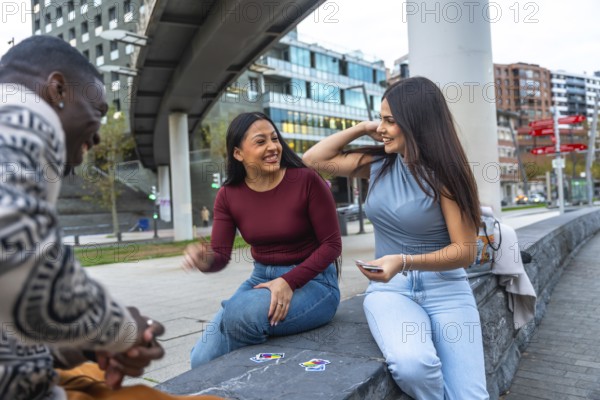 Happy diverse friends sitting in an urban park, playing cards and laughing together, enjoying relaxed outdoor leisure, conversation and casual bonding on a sunny day