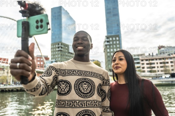 Smiling diverse couple creating content for social media, capturing a selfie video with a smartphone on a vlog stick in an urban setting with modern buildings