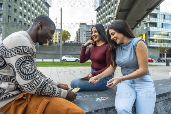 Diverse group of friends sitting outdoors in the city, smiling and laughing while playing cards, enjoying relaxed leisure time, connection and genuine youthful fun