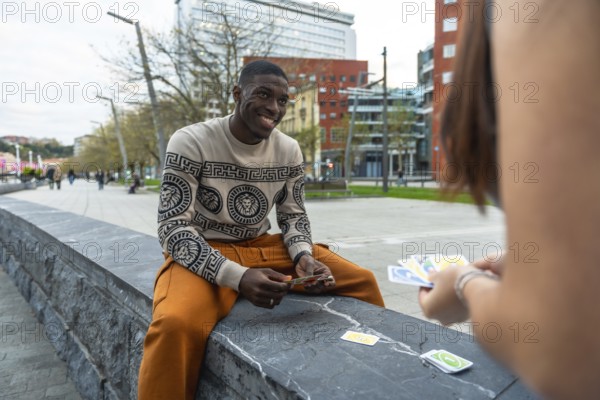 Young man smiling as he and a woman play a competitive card game on a stone bench in a modern urban park, enjoying sunny leisure, friendly competition and connection