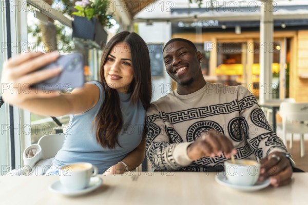 Happy diverse couple smiling while taking a selfie with a smartphone at a coffee shop, enjoying their date and connecting during a relaxed social moment