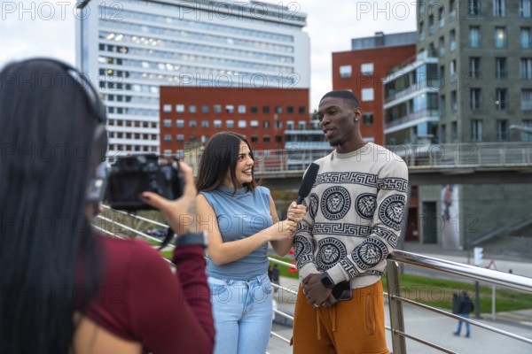 Reporter holding microphone and interviewing a young diverse man on a modern city street, while a camera operator films them for journalism content and media production