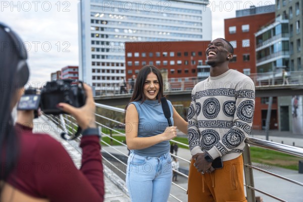 Camerawoman filming an interview with a female reporter holding a microphone and a young cheerful man laughing during a street report on a modern city bridge