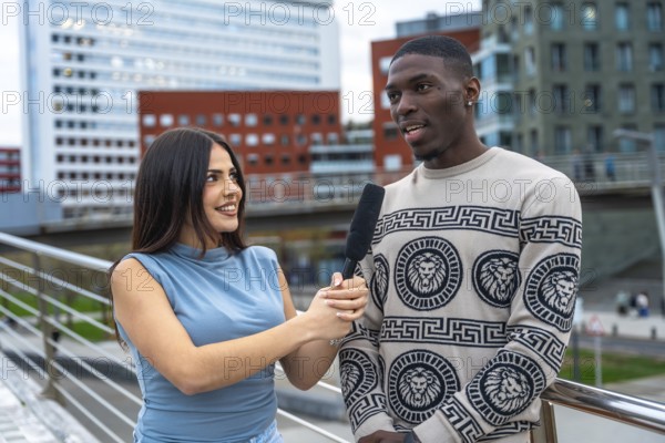 Female journalist interviewing a young black man on a city street, holding a microphone while he shares his thoughts, participating in public opinion gathering