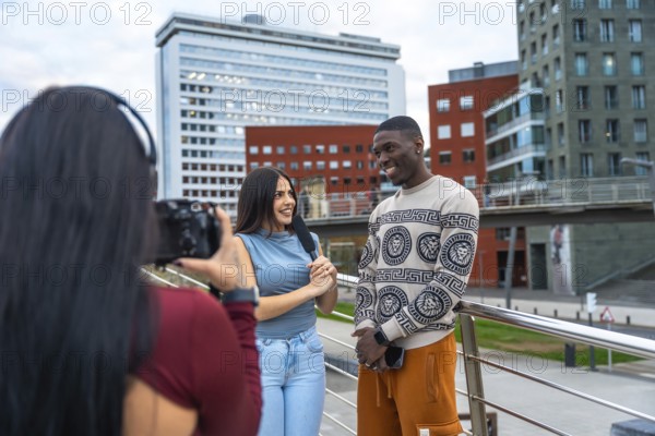 Journalist holding a microphone and talking with a smiling young man while a videographer is filming the scene for a street interview in an urban environment