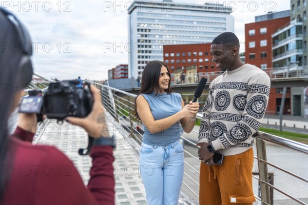 Journalist holding a microphone and interviewing a smiling young man on a modern urban bridge while a third person is filming with a professional camera
