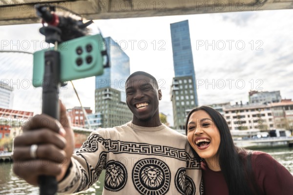 Diverse young couple vlogging together outdoors in a modern city, smiling and laughing as they live stream travel adventures with a smartphone and selfie stick, sharing joy and connection