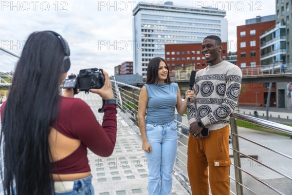 Woman filming an outdoor interview with a young woman holding a microphone to a laughing man, capturing diverse content for vlogging or news in an urban environment
