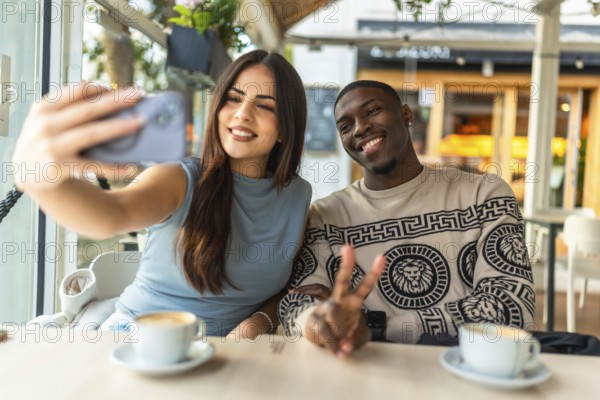 Smiling diverse couple making a peace sign gesture while taking a selfie with a smartphone at a cozy coffee shop, enjoying a beverage and cherishing a fun moment together