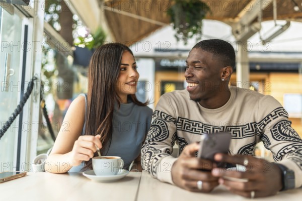 Diverse couple smiling and interacting joyfully, sharing a relaxed moment together at a cafe while looking at a smartphone, symbolizing connection and happiness