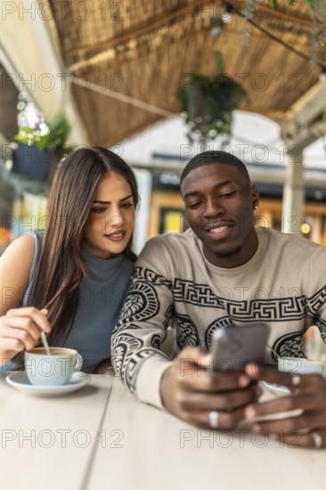 Diverse young woman and man sitting at an outdoor cafe table, smiling and sharing coffee while browsing a smartphone together, enjoying a relaxed digital moment in the city
