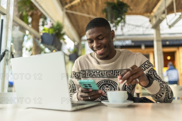 Young black man in a coffee shop using a laptop and smartphone, stirring his coffee while smiling, enjoying freelance work and digital nomad lifestyle