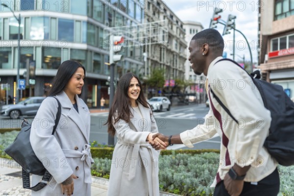 Diverse young adults meeting on a city sidewalk, one woman shaking hands with a man, while another woman stands observing, all smiling during their urban street encounter