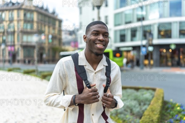 Young black man with a backpack smiling at the camera on a city street, exploring urban architecture and enjoying a confident, casual daytime stroll through downtown scenery