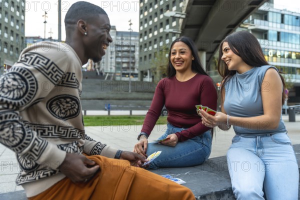 Diverse young friends laughing together while sitting and playing cards outdoors in a modern urban environment, sharing a moment of leisure and entertainment