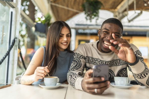 Multiracial couple enjoying a cheerful date, sharing happy moments while viewing media on a mobile phone and drinking coffee in a modern cafeteria setting