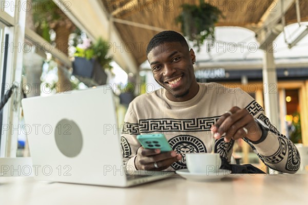Smiling black man sitting at a cafe with laptop and smartphone, stirring coffee while working remotelycasual freelance professional enjoying a relaxed, productive urban workspace