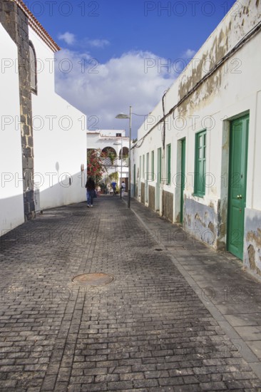 Narrow cobblestone street in Arrecife, Lanzarote, in portrait mode. On the left the back of the church, on the right a row of houses with chipped plaster and green doors, all under a clear blue sky, Arrecife Lanzarote