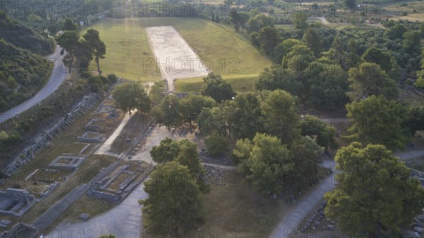 Ancient stadium in the morning backlight, ancient ruins next to a wide meadow, framed by dense tree landscape, drone shot, archaeological site, Ancient Olympia, Peloponnese, Greece
