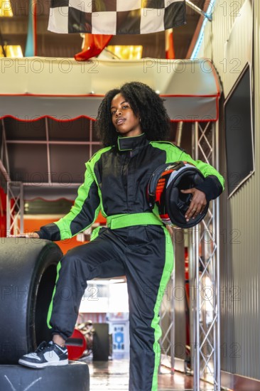 Young black woman standing confidently in a green and black race suit, holding a helmet and resting her foot on a tire, ready for action at an indoor karting track with a checkered flag visible