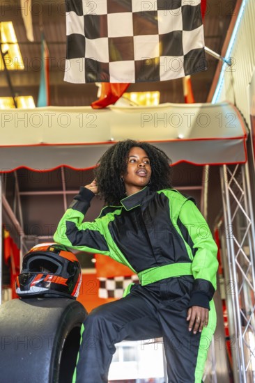 Confident young black woman in a green and black racing jumpsuit and helmet, posing near a kart tire under a checkered flag, preparing for an exciting go karting experience