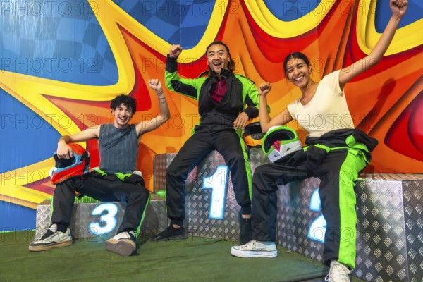 Three young multi ethnic friends on a karting podium, smiling and raising arms in victory while holding helmets after a thrilling race, celebrating teamwork and success