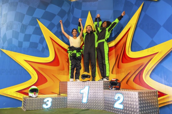 Three friends, dressed in race overalls, raising their arms and celebrating their achievement and success while standing on a winner's podium after a fun karting race
