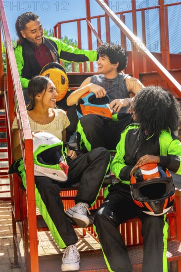 Happy diverse friends group wearing karting suits and helmets, sitting on metal stairs, laughing and enjoying leisure time together after racing go karts