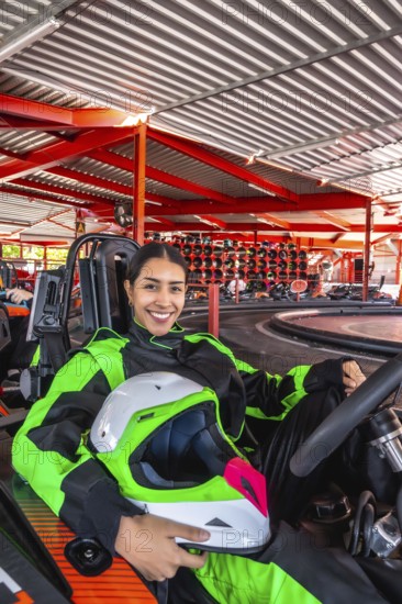Young hispanic woman wearing a racing suit and holding a helmet, sitting in a go kart on the track, smiling at the camera, conveying excitement for the motorsport activity