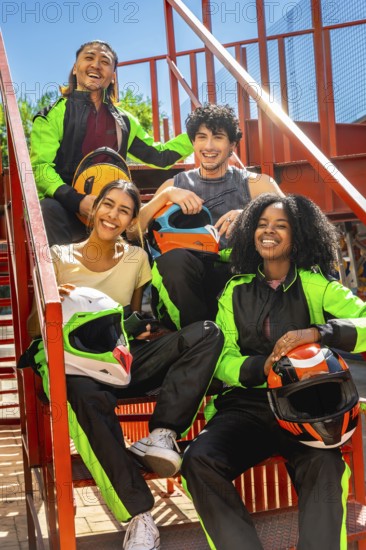 Diverse friends in racing suits holding helmets, smiling and sitting on red metallic stairs outside, celebrating the excitement and fun of karting as a group leisure activity