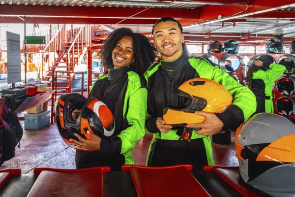 Multiracial young couple smiling at camera in racing suits and helmets, standing on indoor go kart track ready for a fun, excited karting adventure and weekend racing together