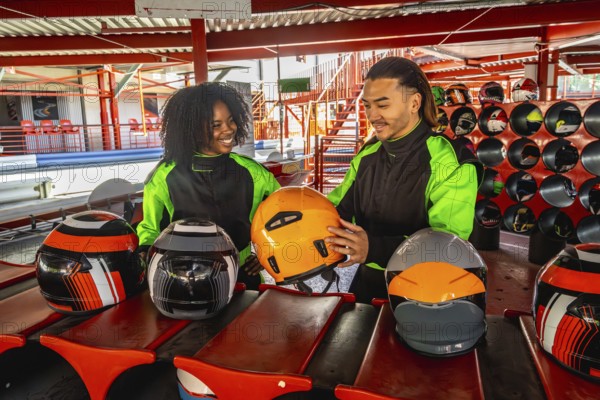 Diverse couple wearing racing suits and choosing helmets, standing on a go kart track, anticipating a fun and thrilling motorsport experience together