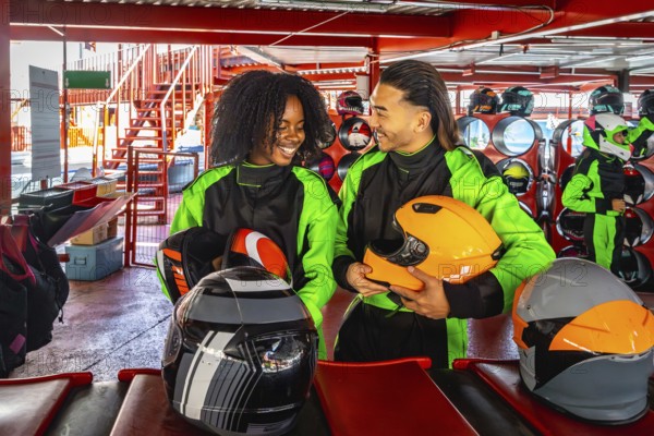 Happy young multiracial couple standing in an indoor karting track area, holding helmets, and looking at each other, enjoying a fun recreational activity together