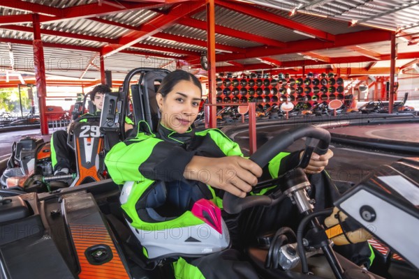 Woman in racing suit grips go kart steering wheel, seated on indoor track ready for competition, focused, helmeted driver exudes determination, speed and weekend motorsport fun