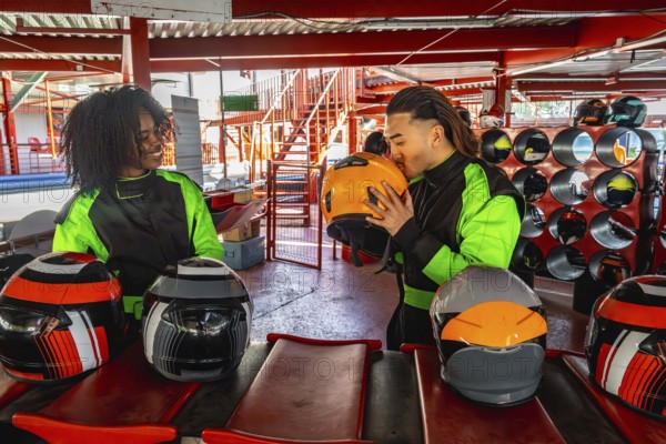 Diverse couple wearing racing suits, one partner kissing a helmet, getting ready for a go karting session at an indoor track, having fun while creating lasting memories together