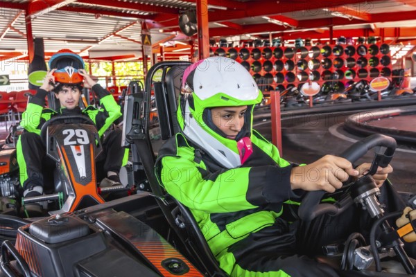 Two young men, one putting on his helmet and another holding a steering wheel, are preparing for a fun karting competition at an indoor go kart racing facility