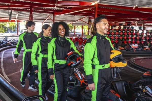 Group of diverse young adults wearing racing suits, carrying helmets, and smiling while walking towards the karts, ready for a fun competition at an indoor karting track