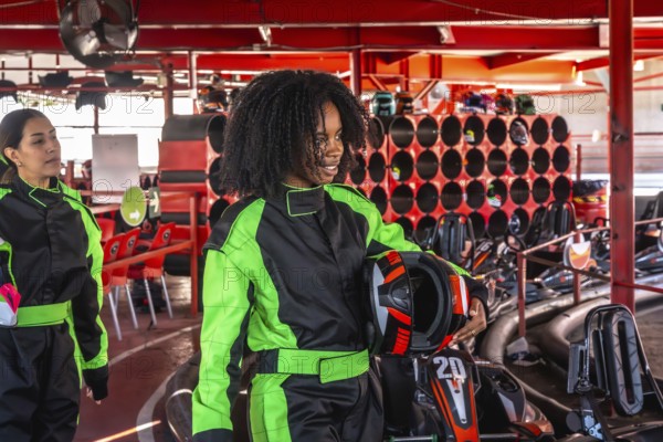 Two cheerful young women in racing suits and holding helmets preparing to race go karts at an indoor track, enjoying a fun day of speed and competition