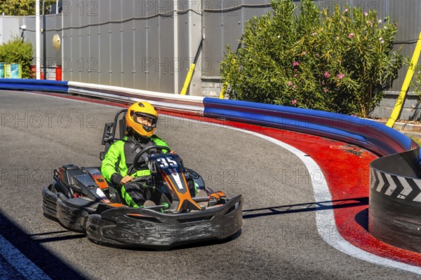 Person wearing a green racing suit and a yellow helmet is driving a go kart around a curve on an outdoor racing track, enjoying the speed and competition involved in motorsport recreation